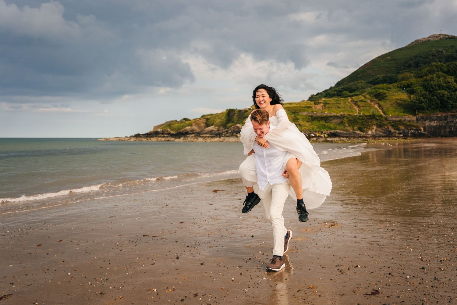 A white-dressed figure runs joyfully along a coastal beach with green hills and stormy clouds in the background. Wedding PHOTOGRAPHER Dublin