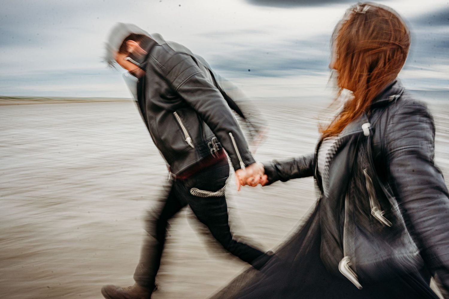 A couple wearing leather jackets hold hands while running on a beach under moody cloudy skies. engagement PHOTOGRAPHER Dublin