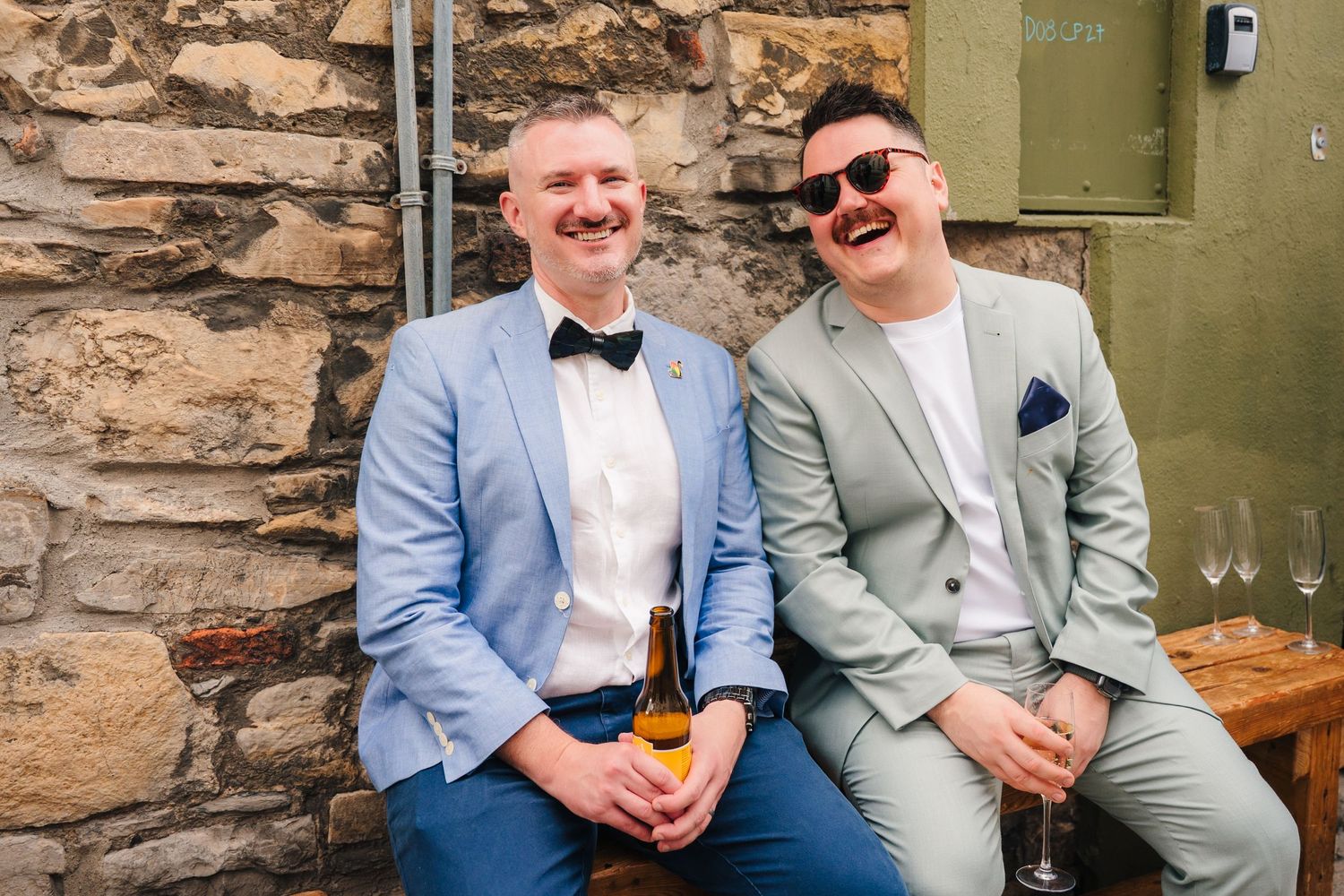 Two formal guests enjoy drinks and conversation while seated by a stone wall at an outdoor event. Same sex wedding PHOTOGRAPHER Dublin