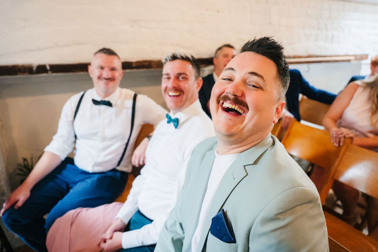 Three men in dark suits laugh together heartily while seated on a bench. Same sex wedding PHOTOGRAPHER Dublin