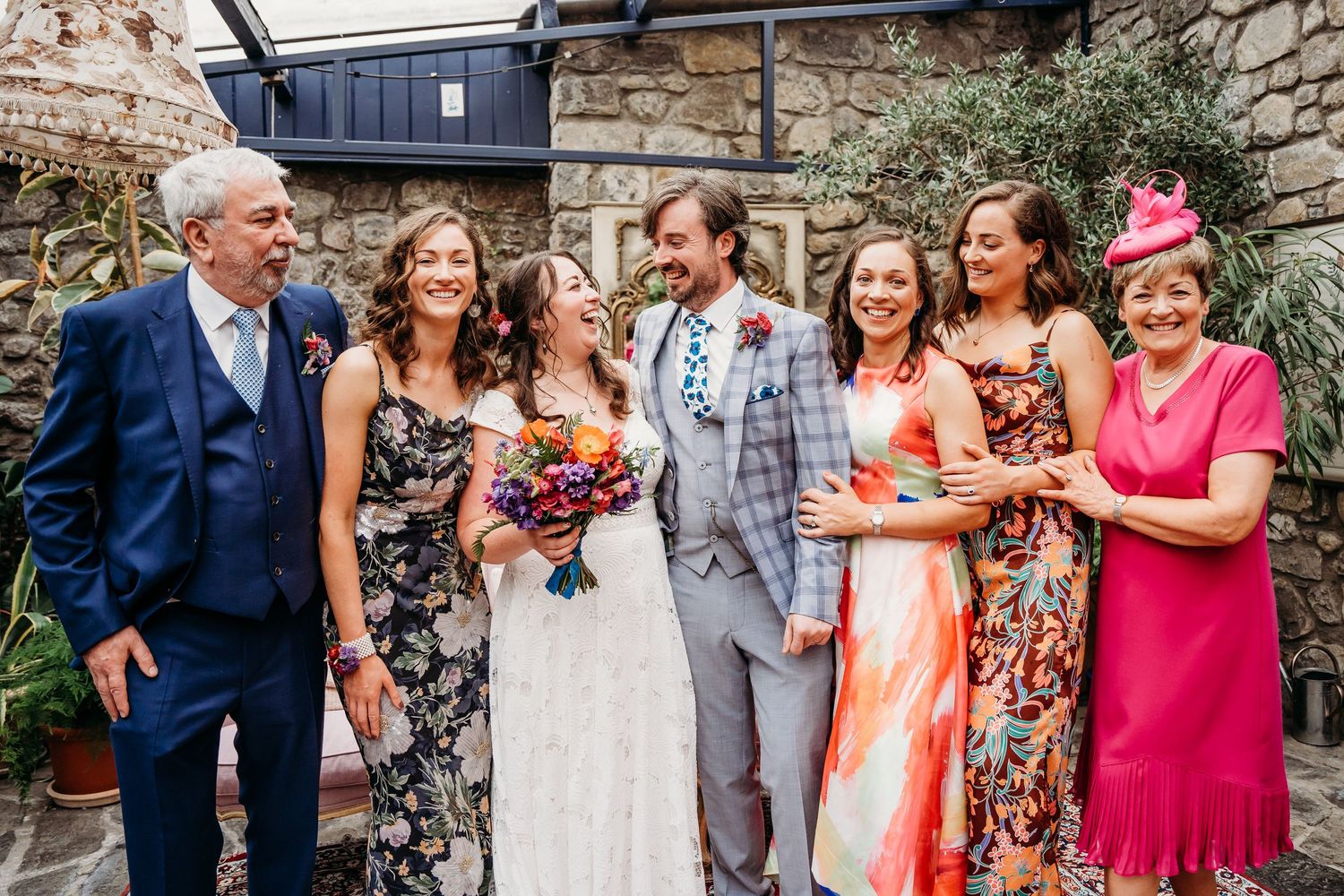 Wedding party in colorful attire posing against stone wall backdrop. Wedding photographer Dublin