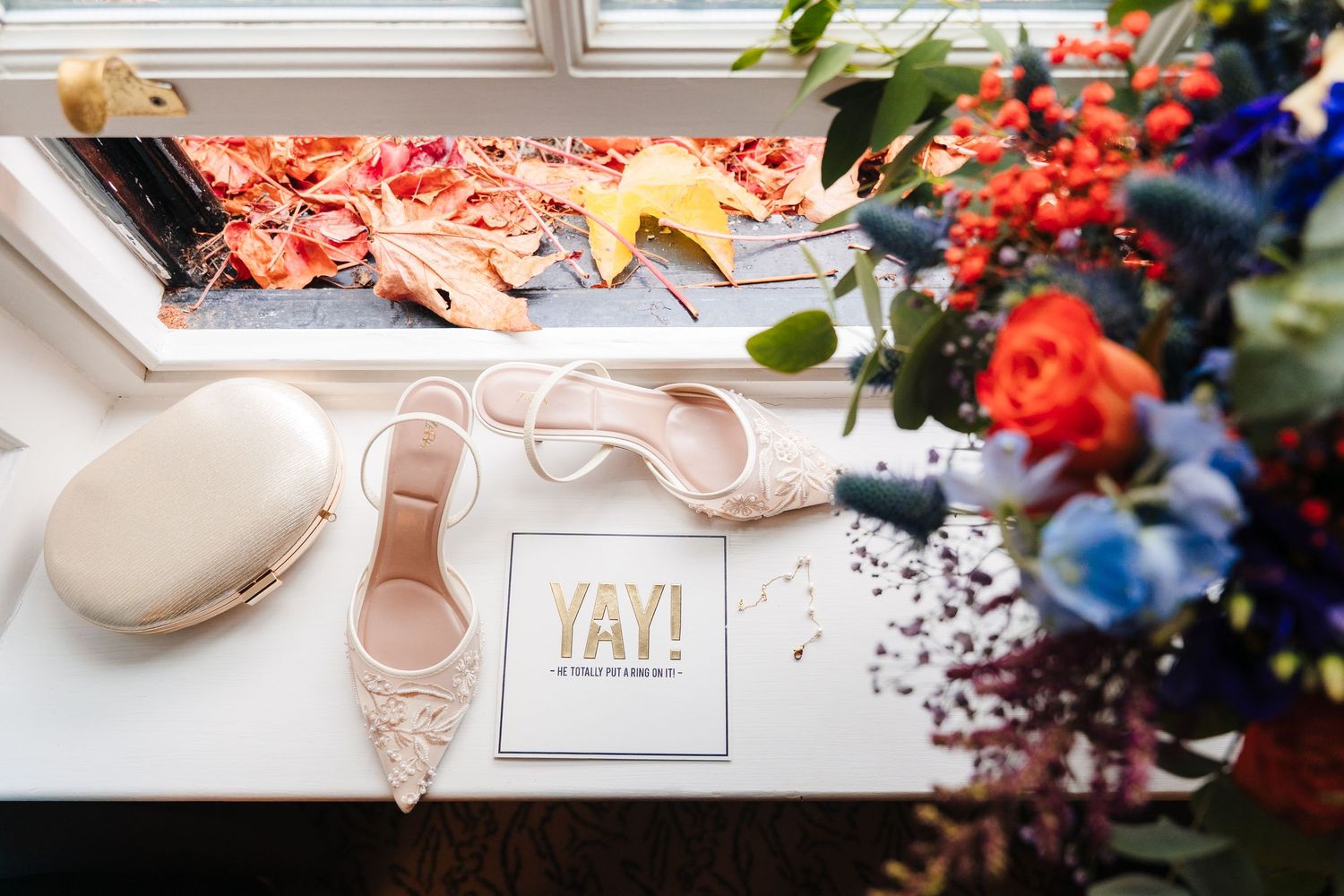 A white wedding shoe sits among colorful floral arrangements and decorative elements on a light surface.