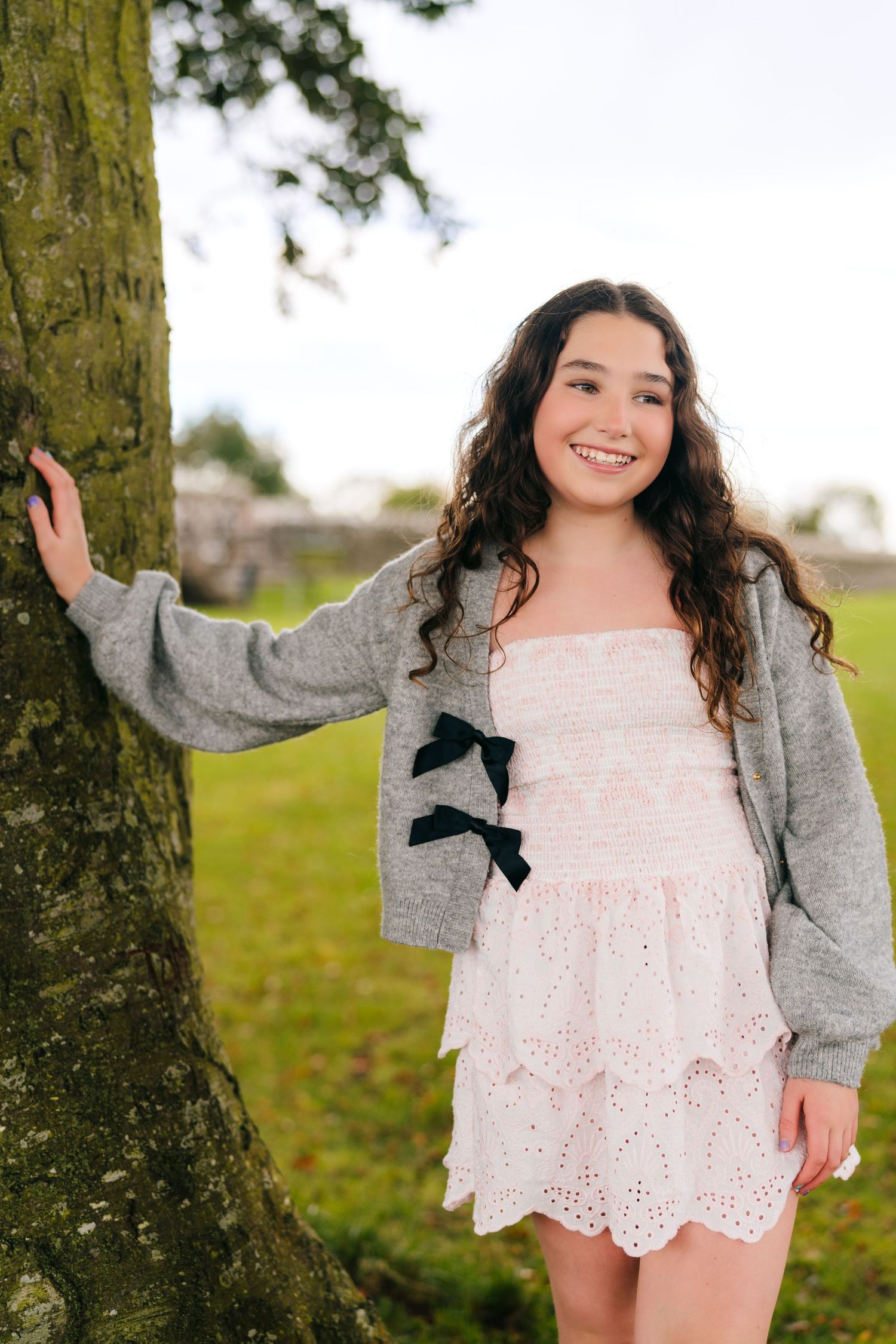 Person in a lace dress and gray cardigan poses playfully near a tree trunk in a park setting. BEctive abbey family photographer dublin
