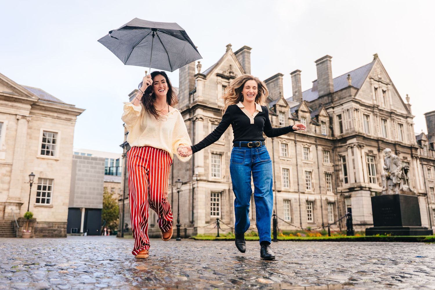 Couple dancing playfully with umbrellas in front of historic stone buildings on a rainy day. professional headshots dublin photographer
