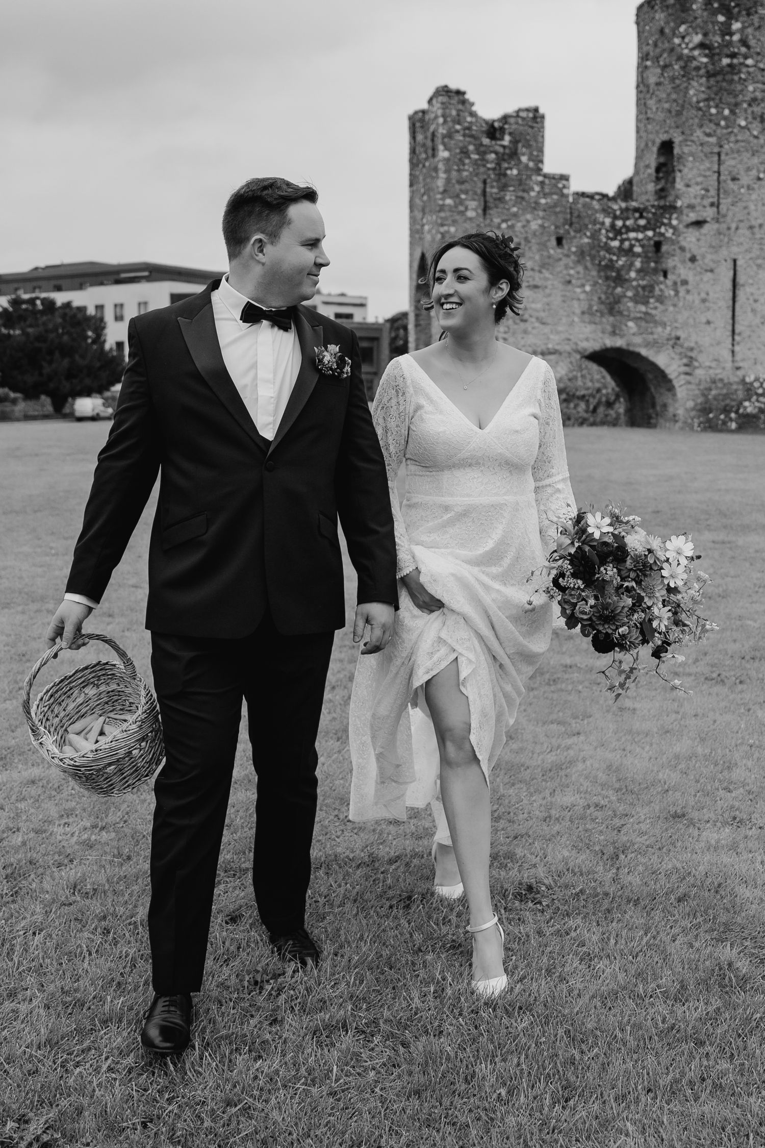 A couple in wedding attire walks hand in hand near ancient castle ruins in black and white. Wedding Photography Trim Castle Ireland