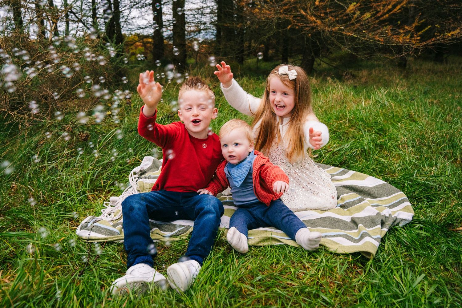 Family Photographer Dublin Ireland Family sitting on blanket playing with bubbles in grassy field near autumn trees.