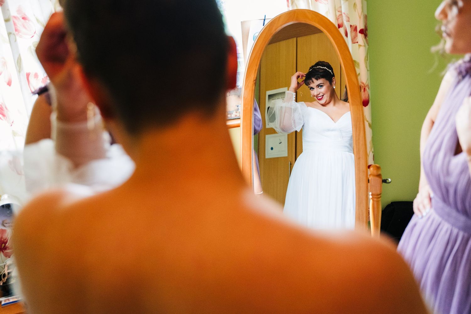 Millhouse slane wedding photographer dublSomeone in a white wedding dress looks in an arched mirror while getting ready with others in view.