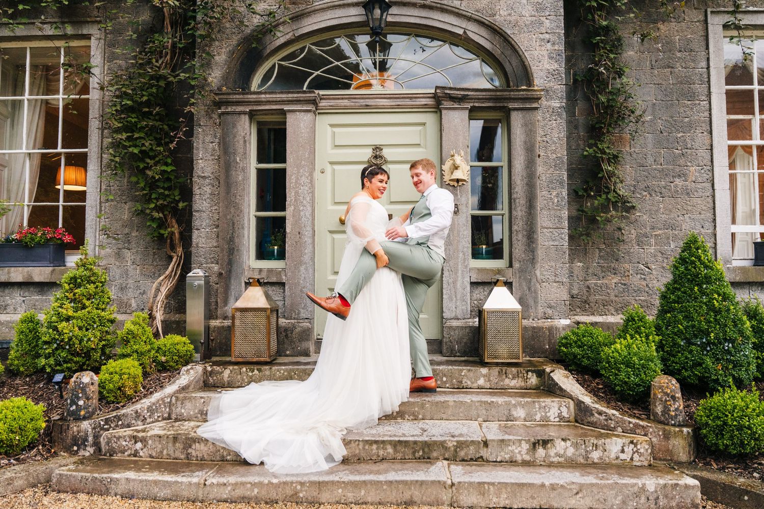 Millhouse slane wedding photographerNewlyweds embrace on grand stone steps of a historic mansion with ivy-covered walls and topiary gardens.