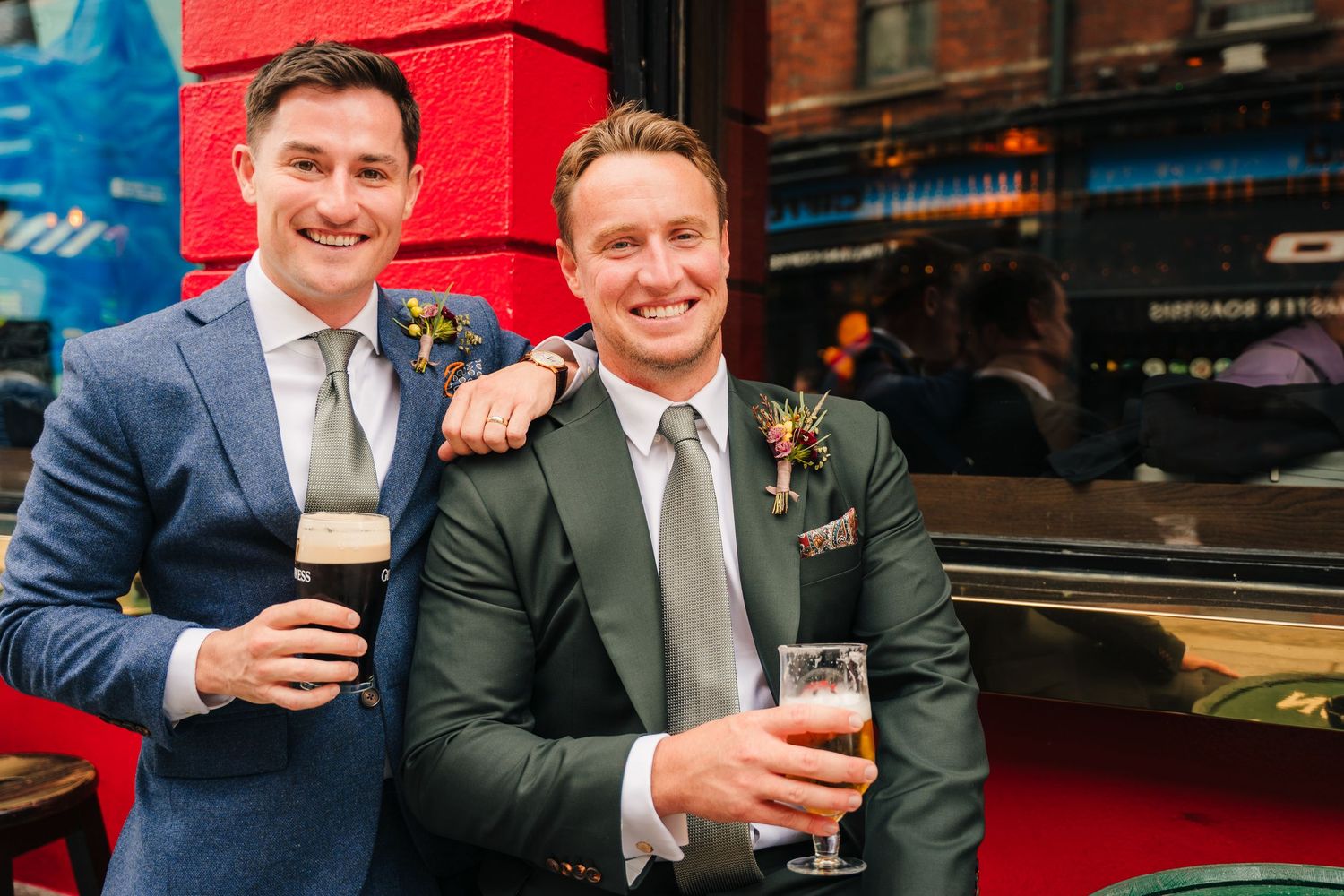 Two people in business attire smile and clink beer glasses in a dimly lit bar setting. Same sex wedding PHOTOGRAPHER Dublin