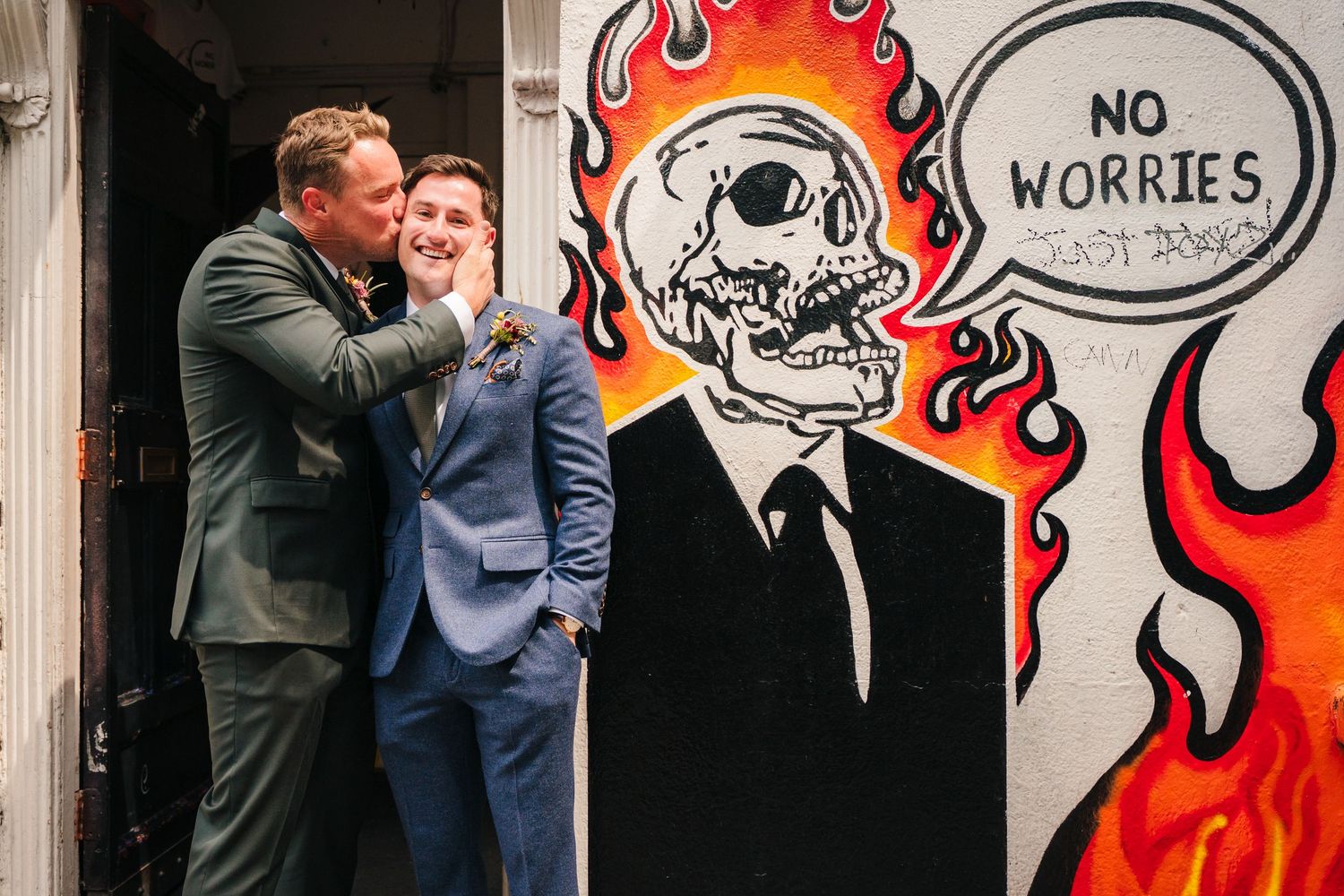 Two people share a joyful moment next to vibrant street art featuring a flaming skull with 'No Worries' text. Same sex wedding PHOTOGRAPHER