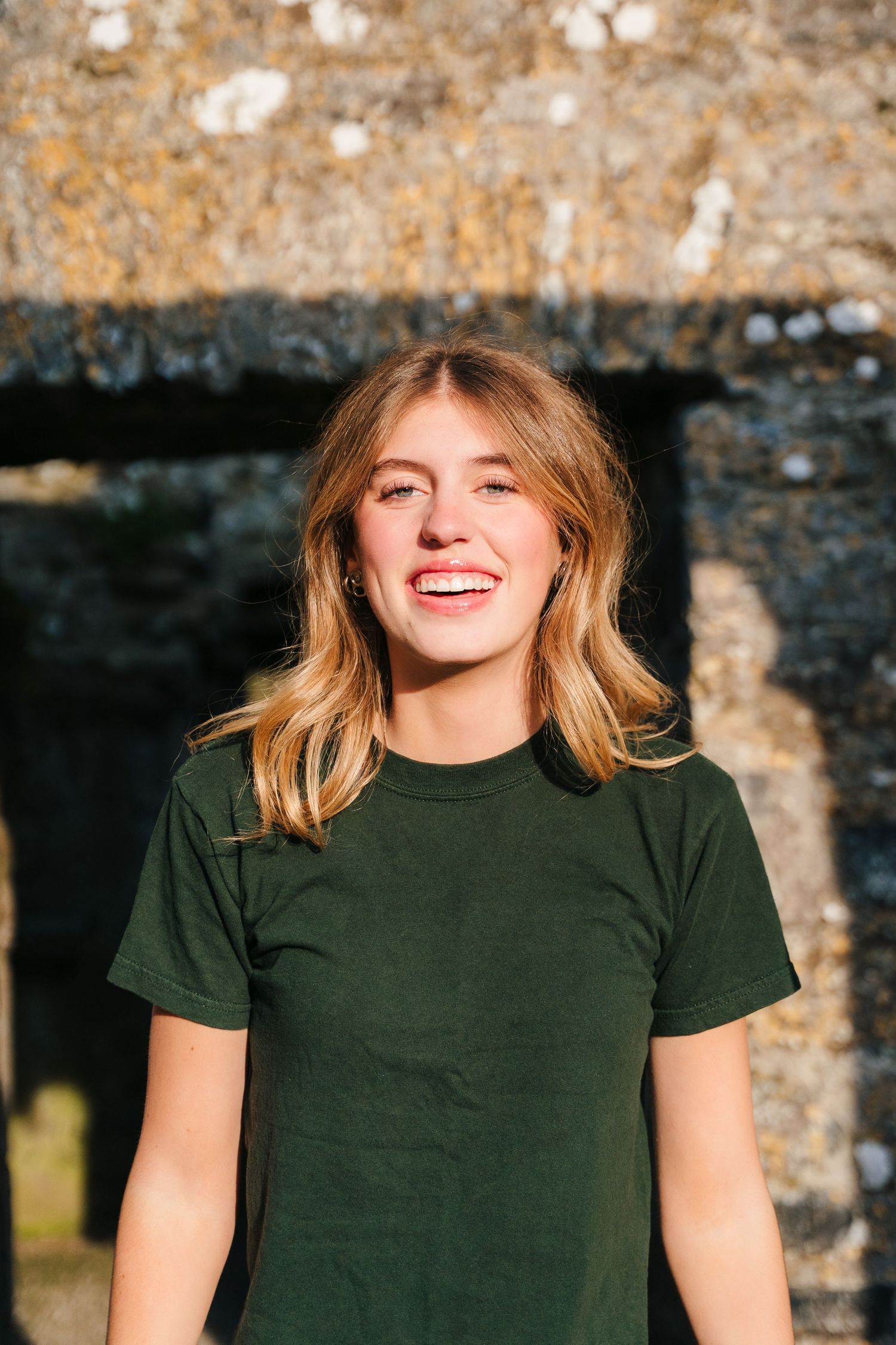 A smiling person with wavy blonde hair wearing a dark green t-shirt stands against a stone wall in sunlight. Photographer Ireland dublin