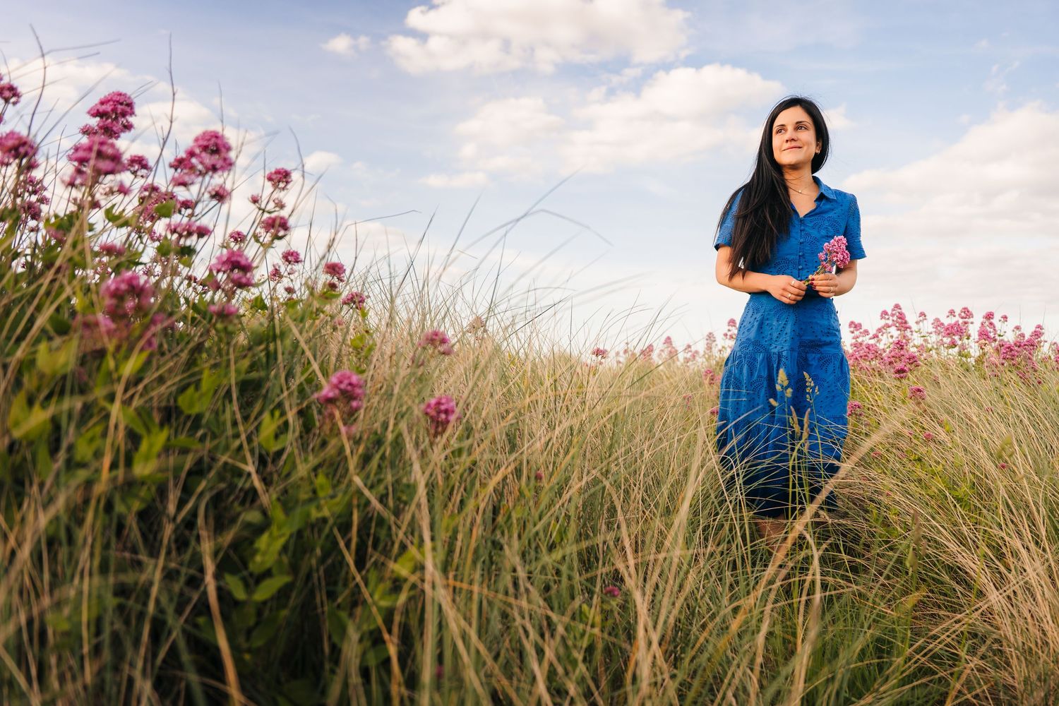 Woman in blue dress standing in tall grass field with pink wildflowers under a partly cloudy sky.