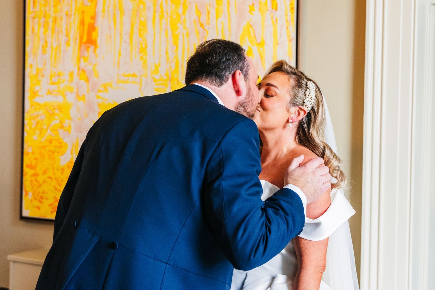 Wedding couple embracing and smiling in front of vibrant yellow artwork in an elegant room. Virginia Park Lodge Wedding Photographer Dublin