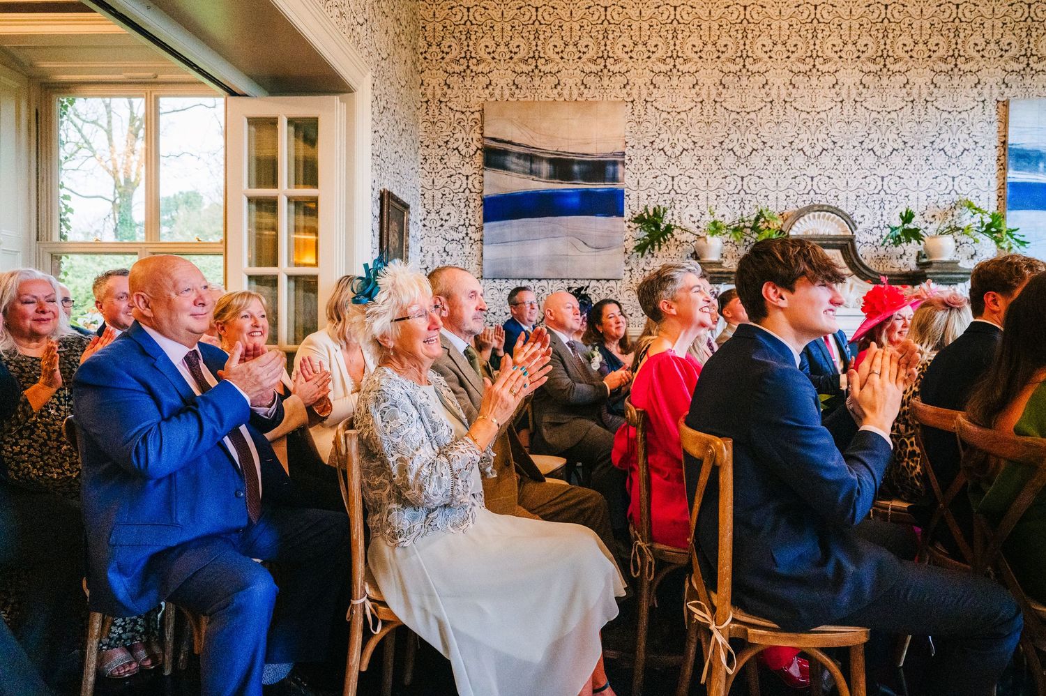 Wedding guests seated in rows applaud during a ceremony in a room with patterned wallpaper.  Virginia Park Lodge Wedding Photographer Cavan