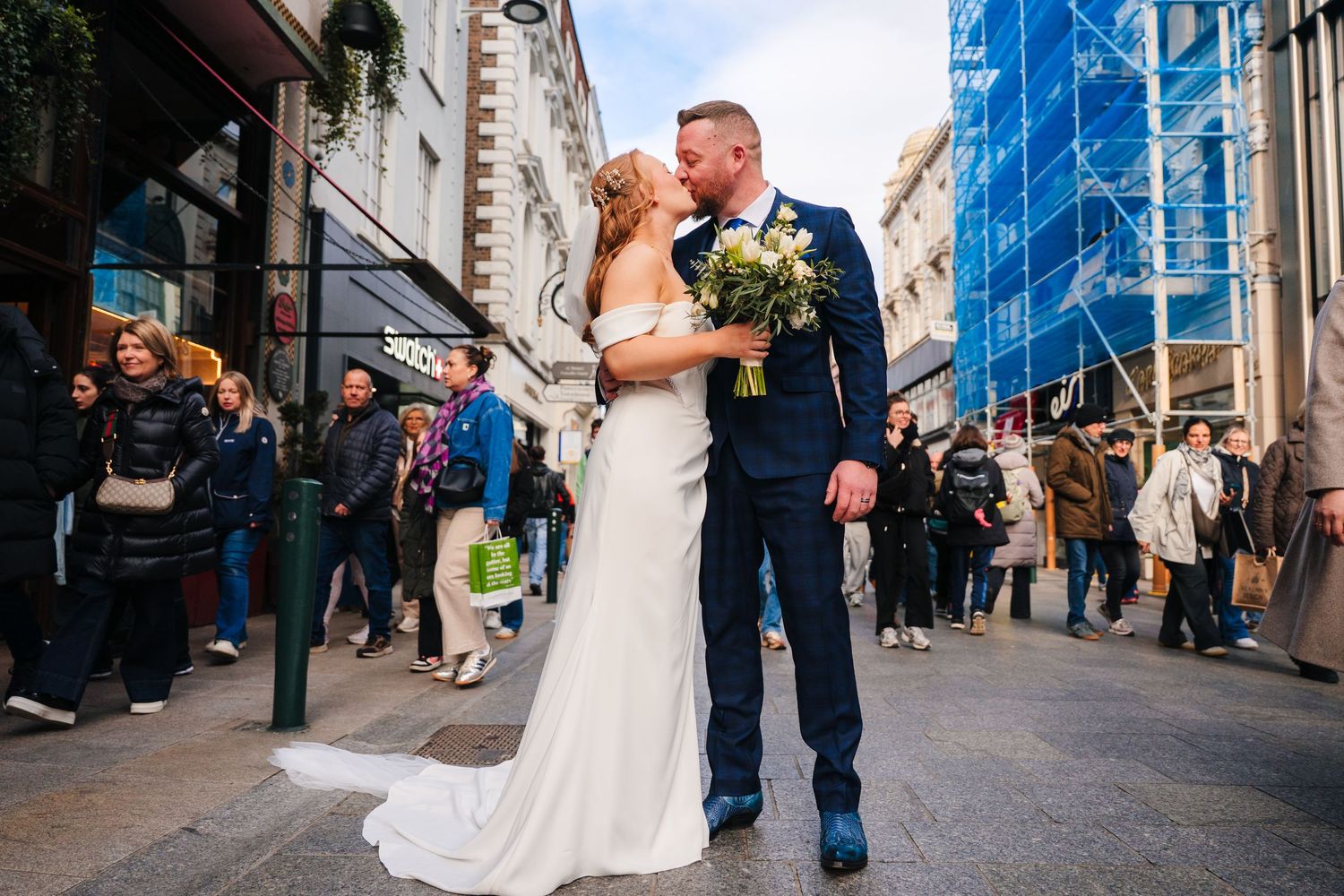 A newlywed couple shares a romantic moment on a busy city street while onlookers gather around them. Dublin wedding photogrAPher Bewleys Gra