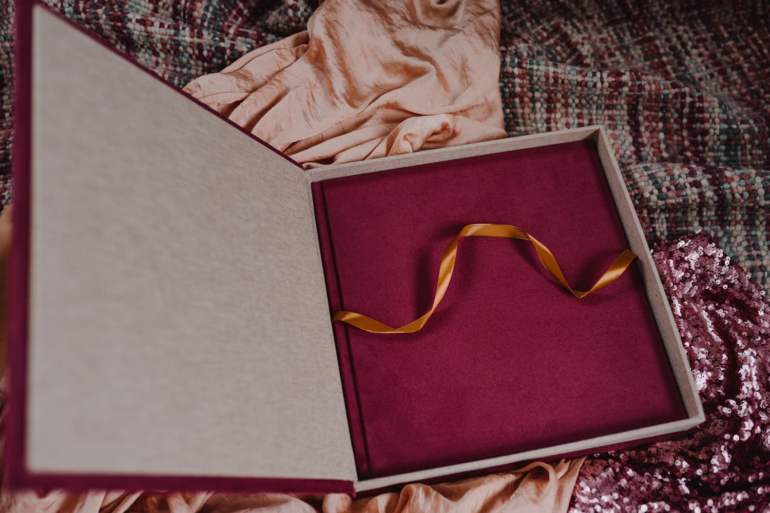 An open gift box with burgundy velvet interior and gold ribbon on textured fabric background.