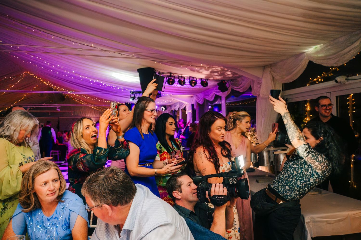 Energetic crowd celebrates at a wedding reception under purple-lit tent with string lights and decorations. Wedding photographer Dublin