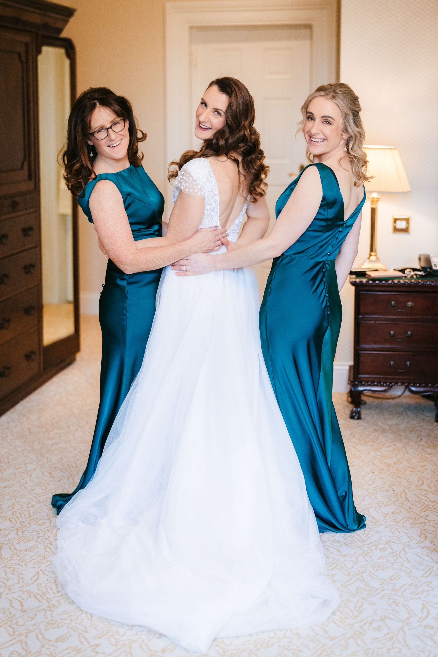 Bridesmaids in teal dresses help adjust the bride's white gown in an elegant indoor setting.