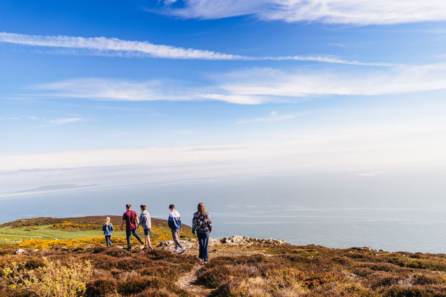 Hikers trek along a scenic coastal trail overlooking a misty ocean under bright blue skies and wispy clouds. Photographer dublin ireland