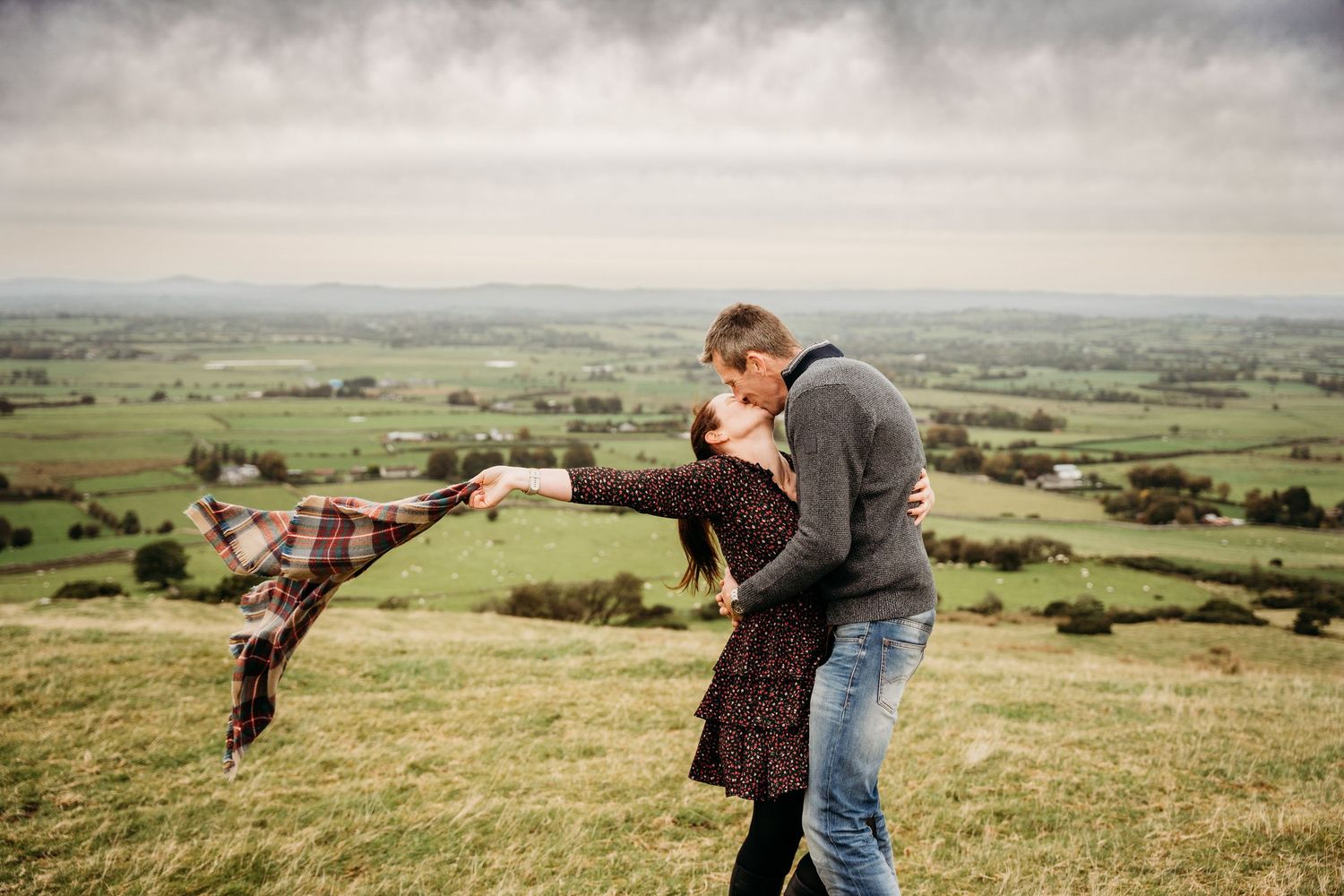 A romantic couple embraces on a hilltop overlooking a vast green countryside landscape on an overcast day. Photographer dublin ireland