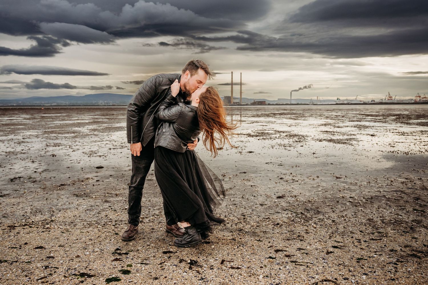 A romantic couple embraces at sunset on a moody beach landscape with dramatic stormy clouds overhead.