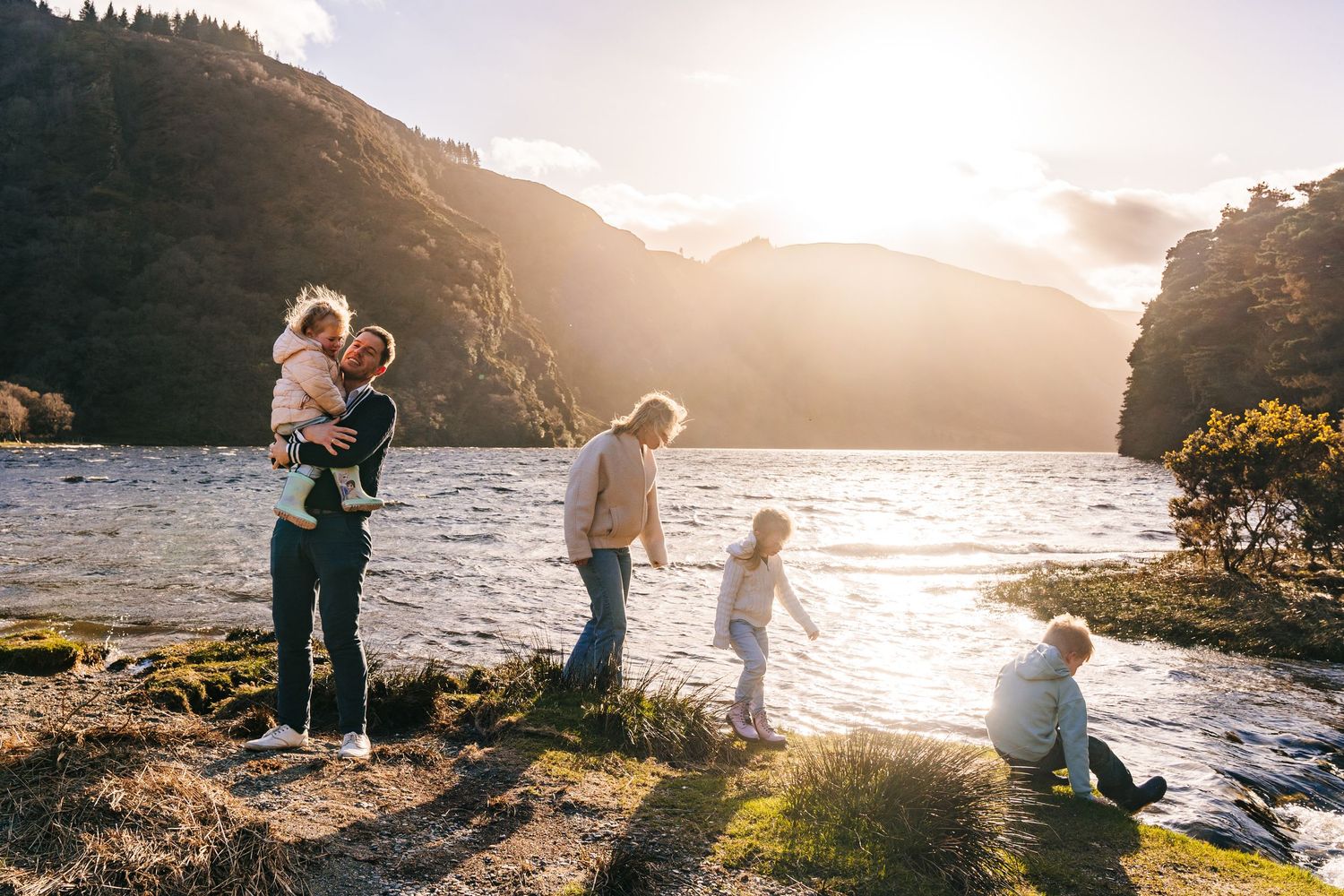 A family enjoys a golden sunset by a scenic lake surrounded by mountains and cliffs. family PHOTOGRAPHER Dublin