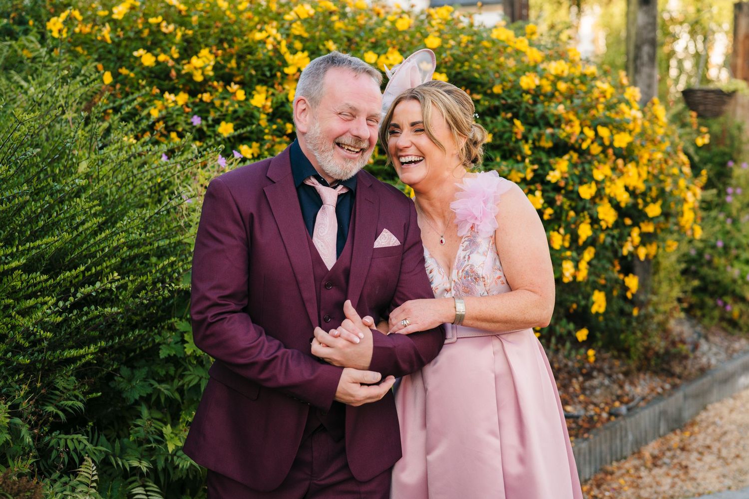 A couple shares a joyful moment together in front of blooming yellow flowers wearing burgundy and blush formal attire. Wedding PHOTOGRAPHER