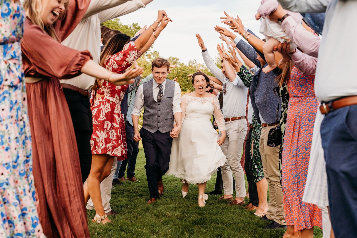 Wedding guests form a cheerful tunnel outdoors as a couple walks beneath raised hands during outdoor celebration. Wedding PHOTOGRAPHER Dubli