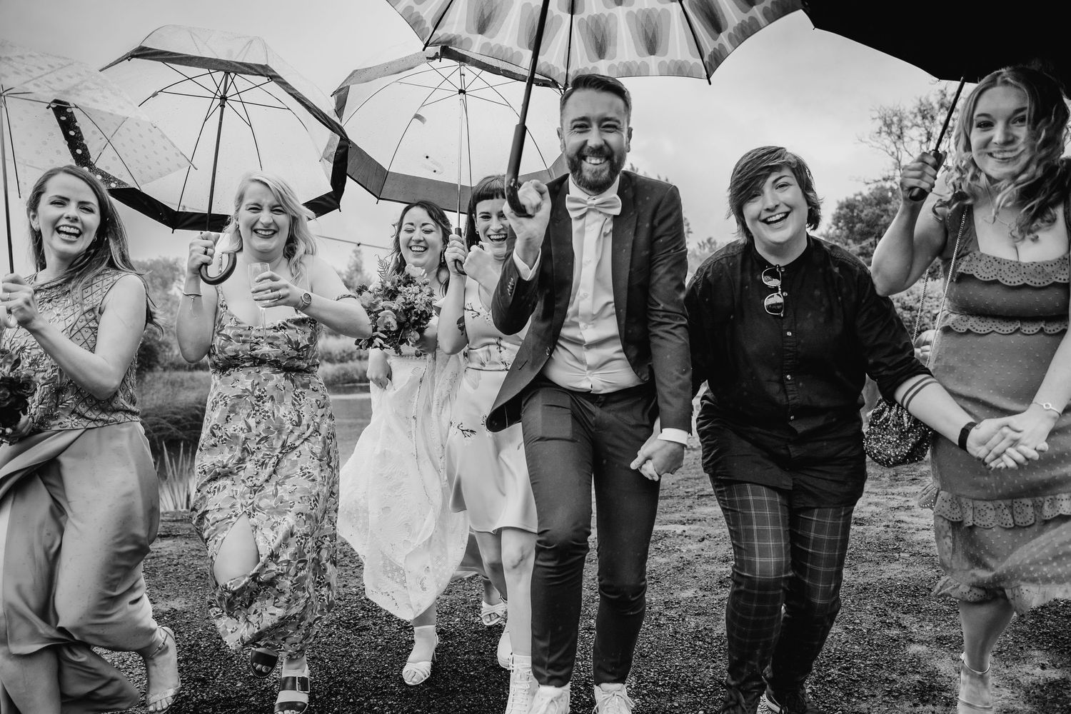 A celebratory group runs through the rain with umbrellas while laughing and jumping in black and white photography. Wedding PHOTOGRAPHER Dub