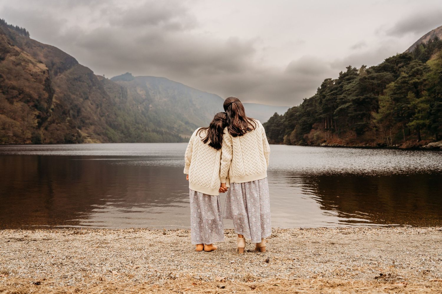 Two people in white outfits standing on a shoreline overlooking a scenic mountain lake on a cloudy day.