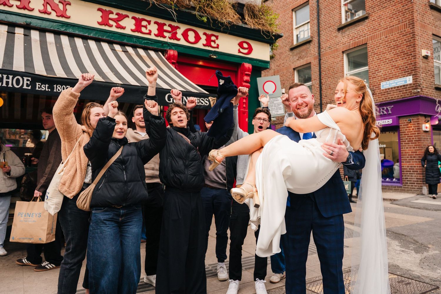 A joyful celebration outside a brick building with a group cheering as someone is lifted up in a celebratory pose. Dublin wedding photogrAPh