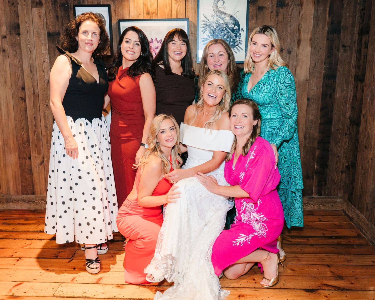 Women in colorful dresses gather for a group photo at a wedding celebration with wooden wall backdrop. The Dean Dublin Wedding Photographer