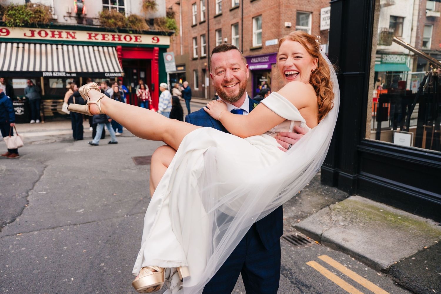 Groom carrying bride in white dress on a cobblestone street outside John kehoe pub in Dublin.