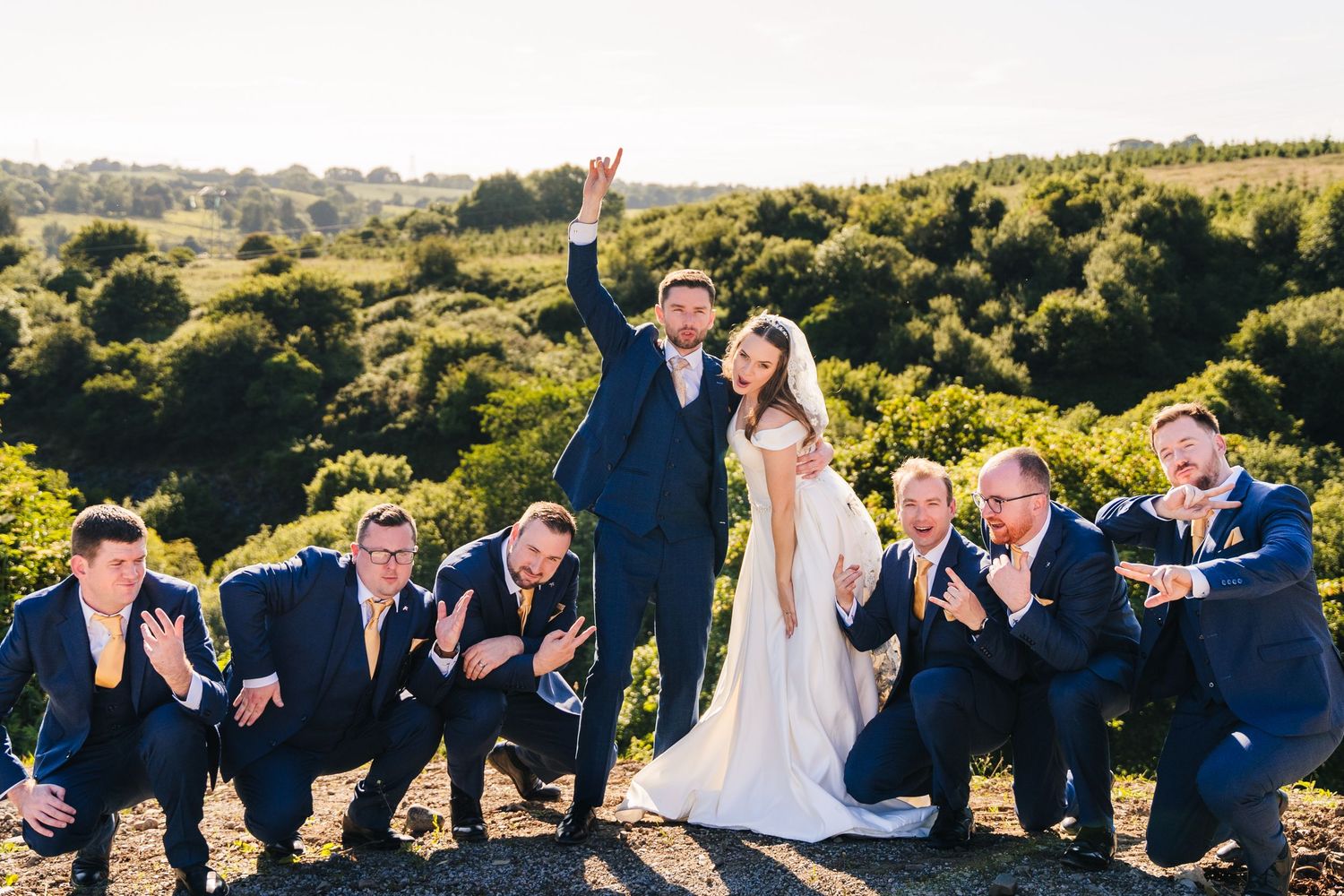 Wedding party in formal navy suits and white gown pose playfully together on grassy hilltop at sunset. Wedding PHOTOGRAPHER Dublin