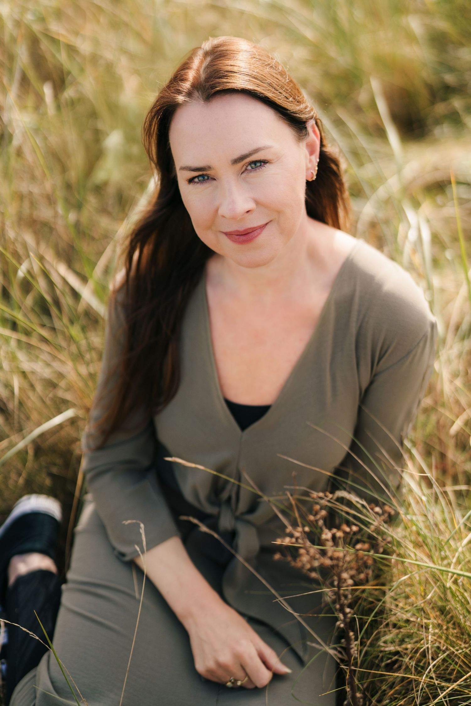 Woman in olive colored top sitting in tall grass on a sunny day during an outdoor portrait session. PHOTOGRAPHER Dublin ireland