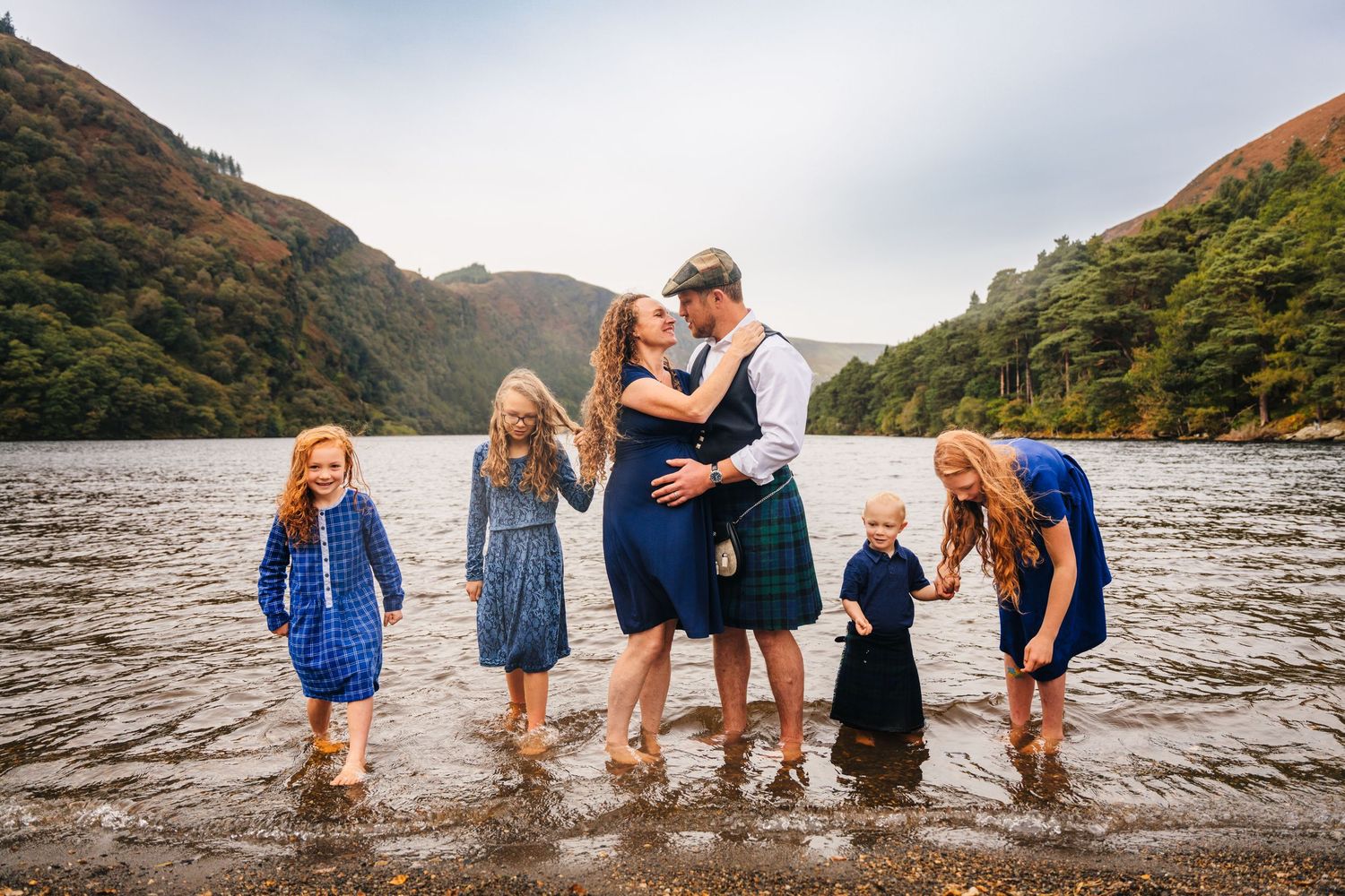 A row of figures stand along the shoreline of a mountain lake beneath cloudy skies. family PHOTOGRAPHER Dublin