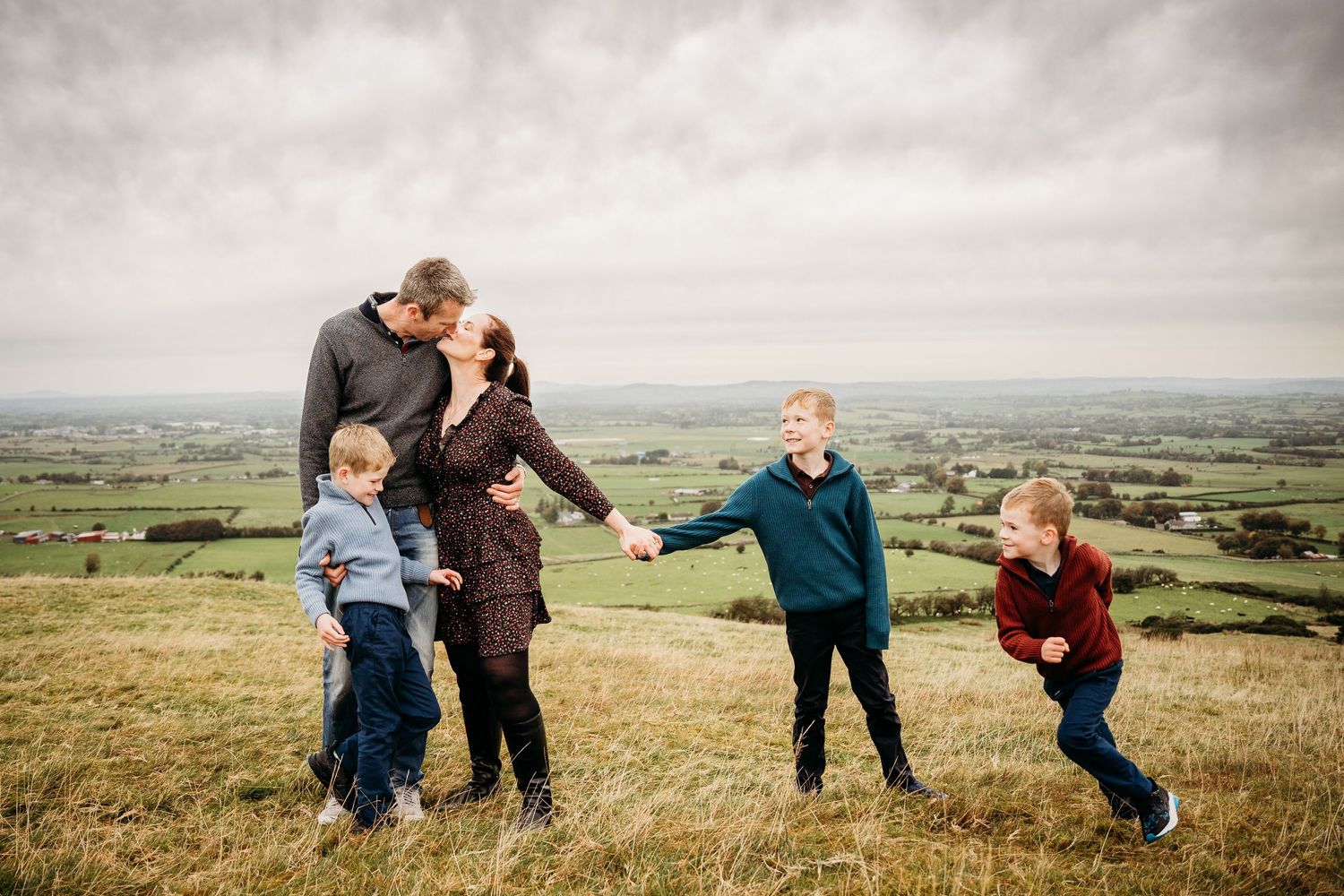 A joyful group shares a playful moment together on a hilltop overlooking vast green countryside in the distance. family PHOTOGRAPHER Dublin