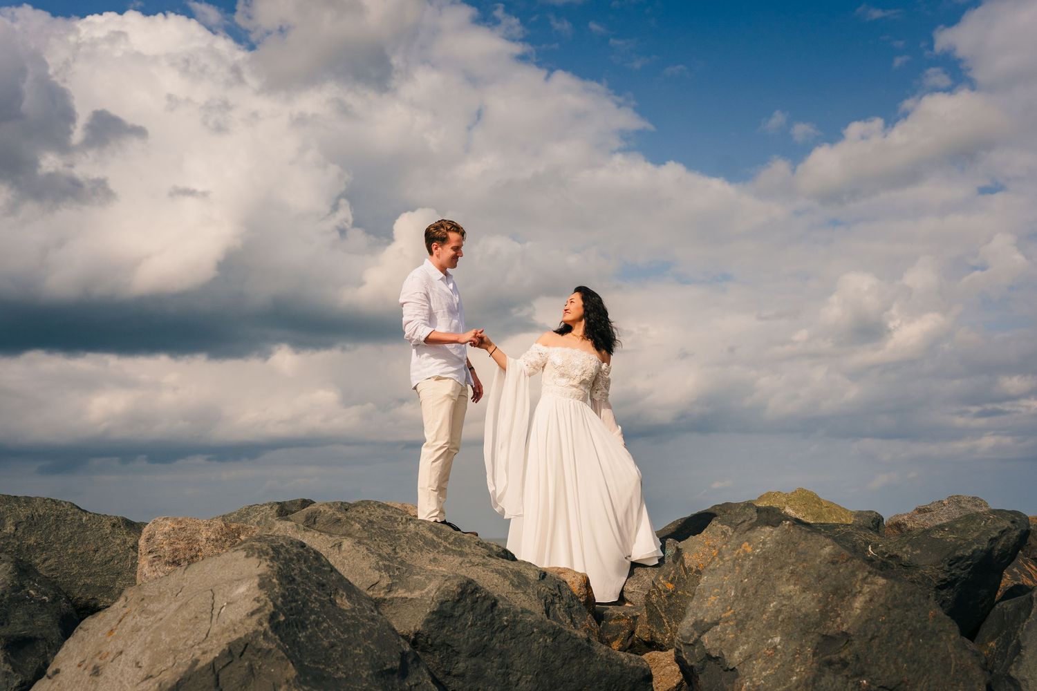 A couple in white attire stands atop rocky coastal terrain against a dramatic cloudy sky during their romantic photoshoot.