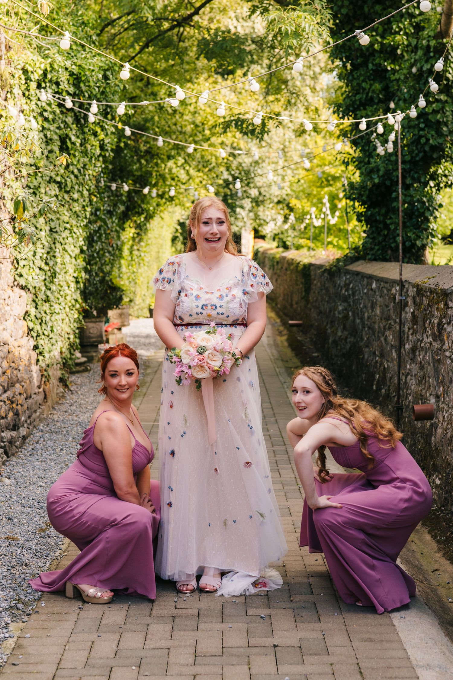 Wedding party poses on brick walkway with floral decor and string lights in garden setting. . Village Barn Tyrrellspass wedding photographer