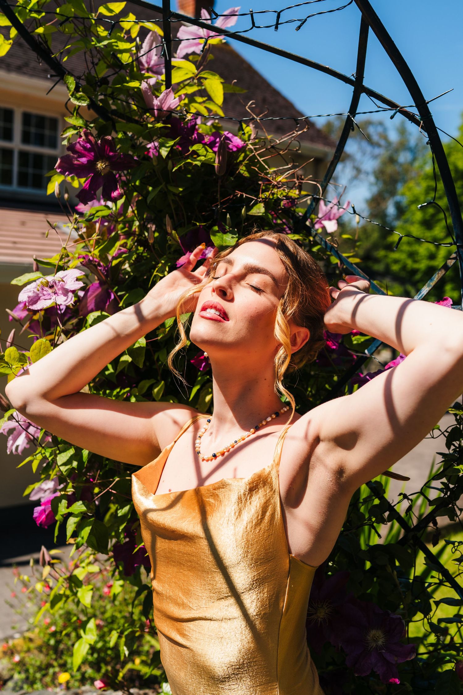 Poulaphouca Wedding photographer wA person in a gold dress poses dramatically against purple flowers and greenery in a sunny garden setting.