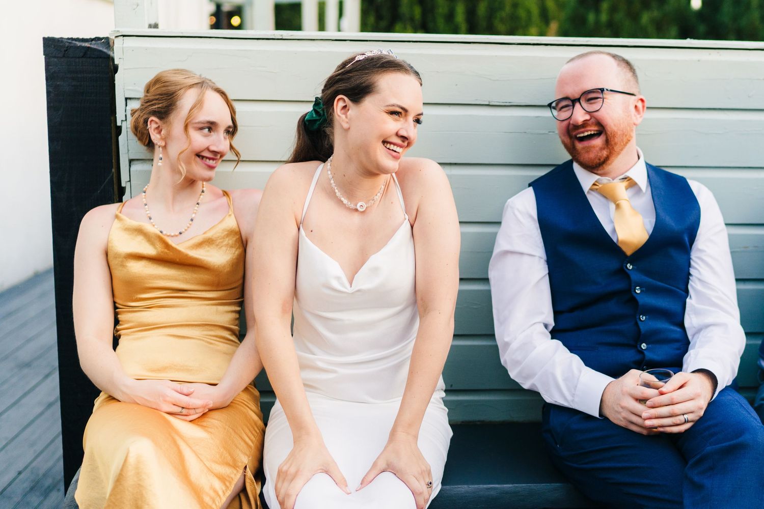 Three wedding attendees share a candid laughing moment while sitting on outdoor bench.
