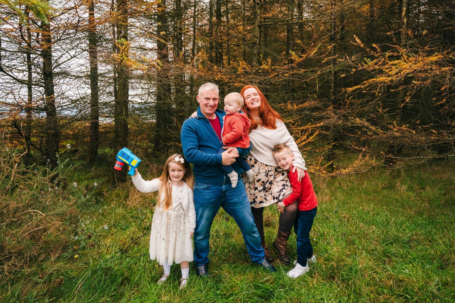 Happy family poses together on grass with autumn forest backdrop.