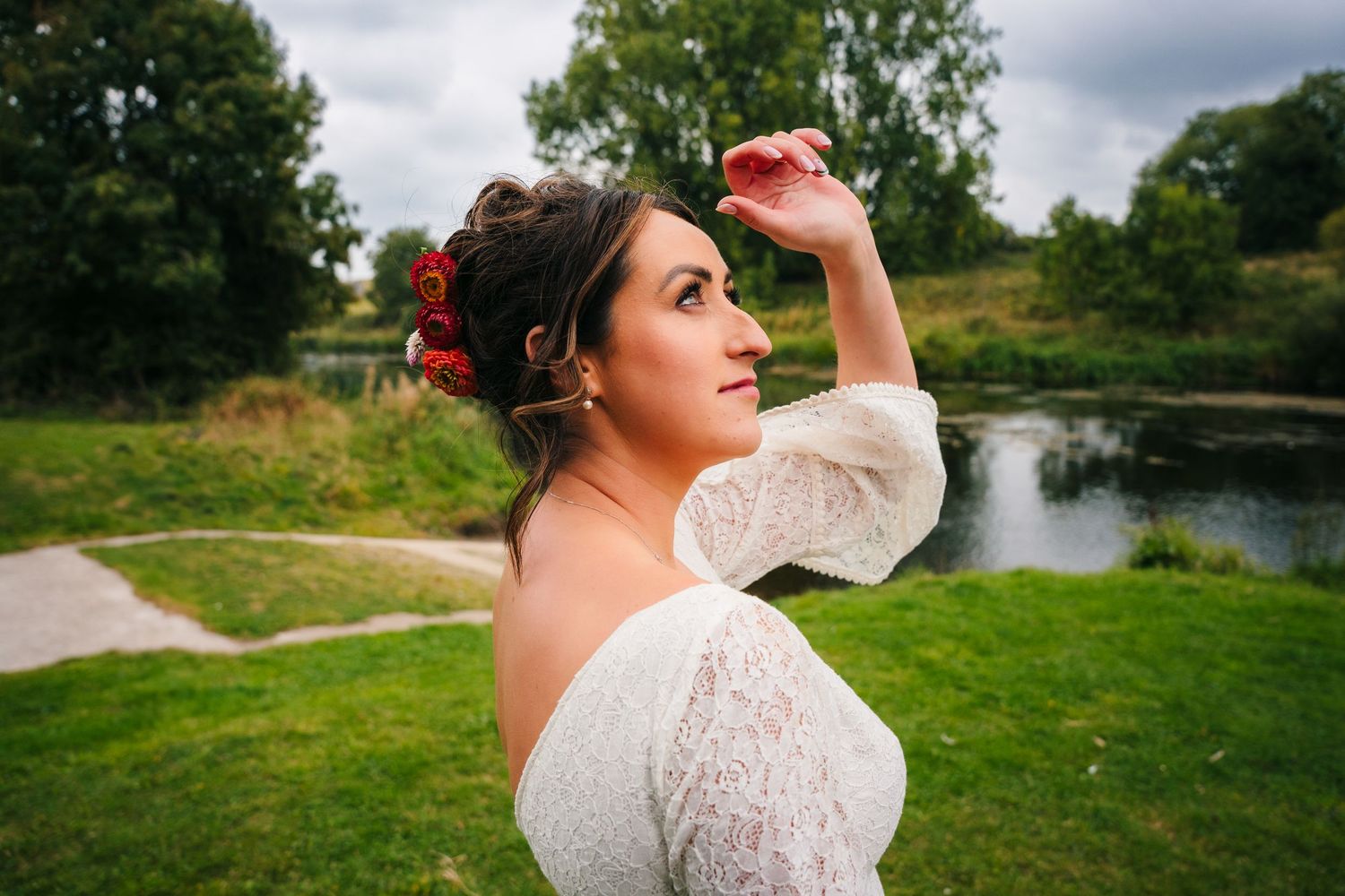 A bride in a lace dress poses near a pond on a grassy lawn with cloudy skies in the background. Wedding Photography Trim Castle Ireland