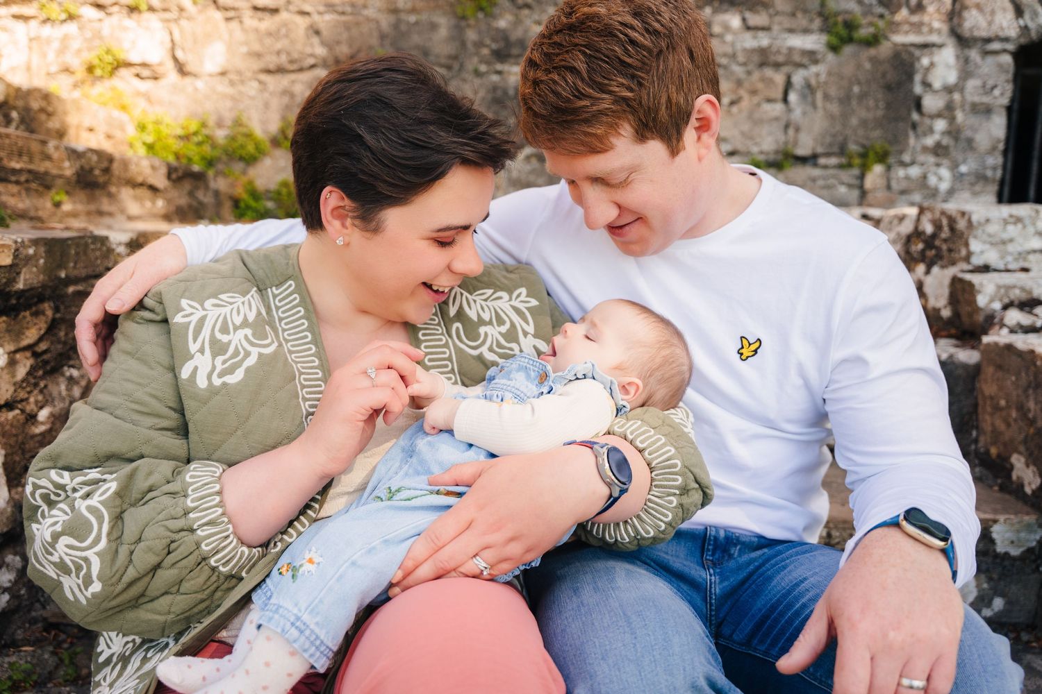 Family PHOTOGRAPHER Dublin A tender family moment captured outdoors as they cradle their newborn baby against a stone wall background.
