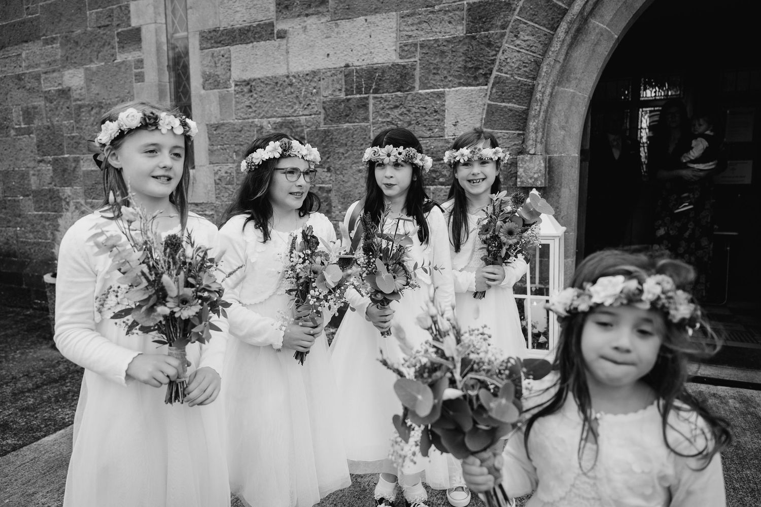 Wedding party wearing floral crowns and white dresses pose together in a joyful outdoor setting.