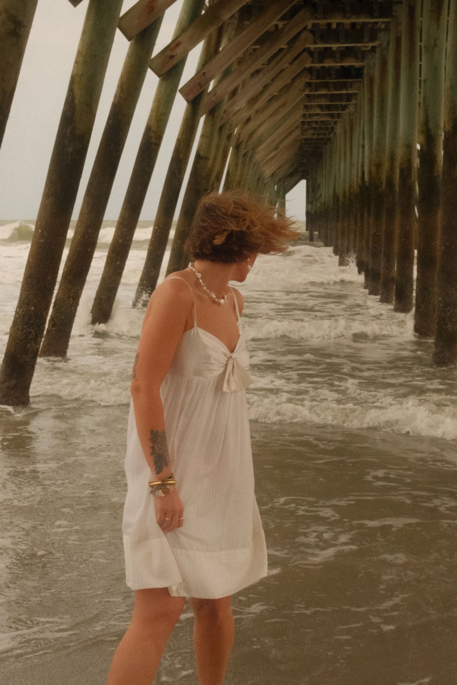 Person in white dress walks along misty beach under wooden pier posts while waves roll in at sunset.