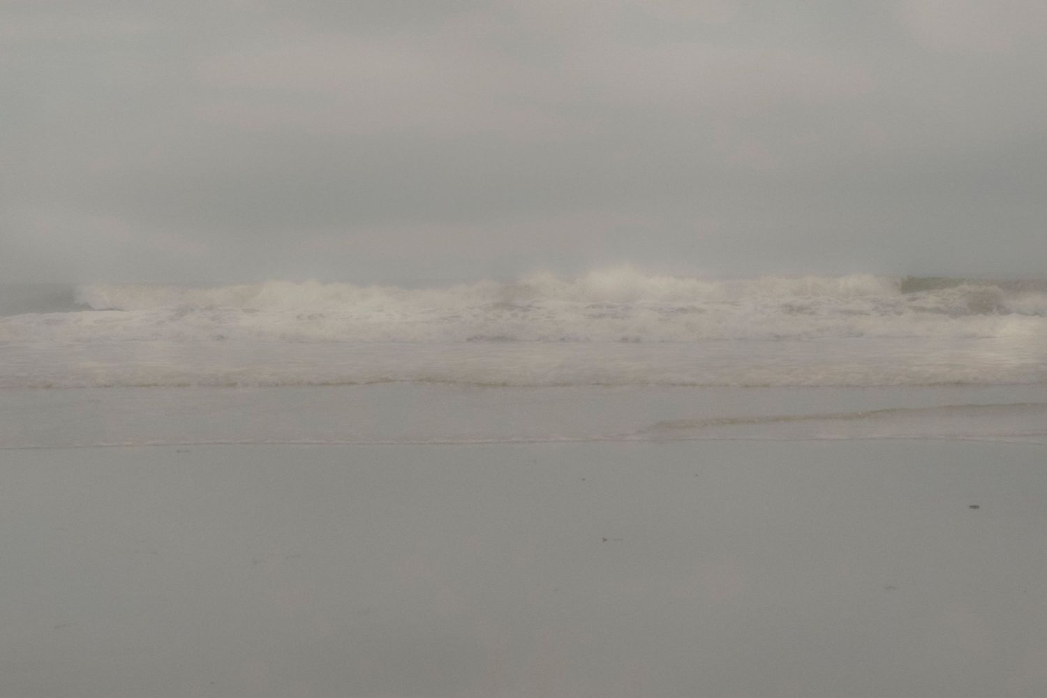 Large waves crash onto a foggy beach shore on an overcast gray day with white foamy surf.