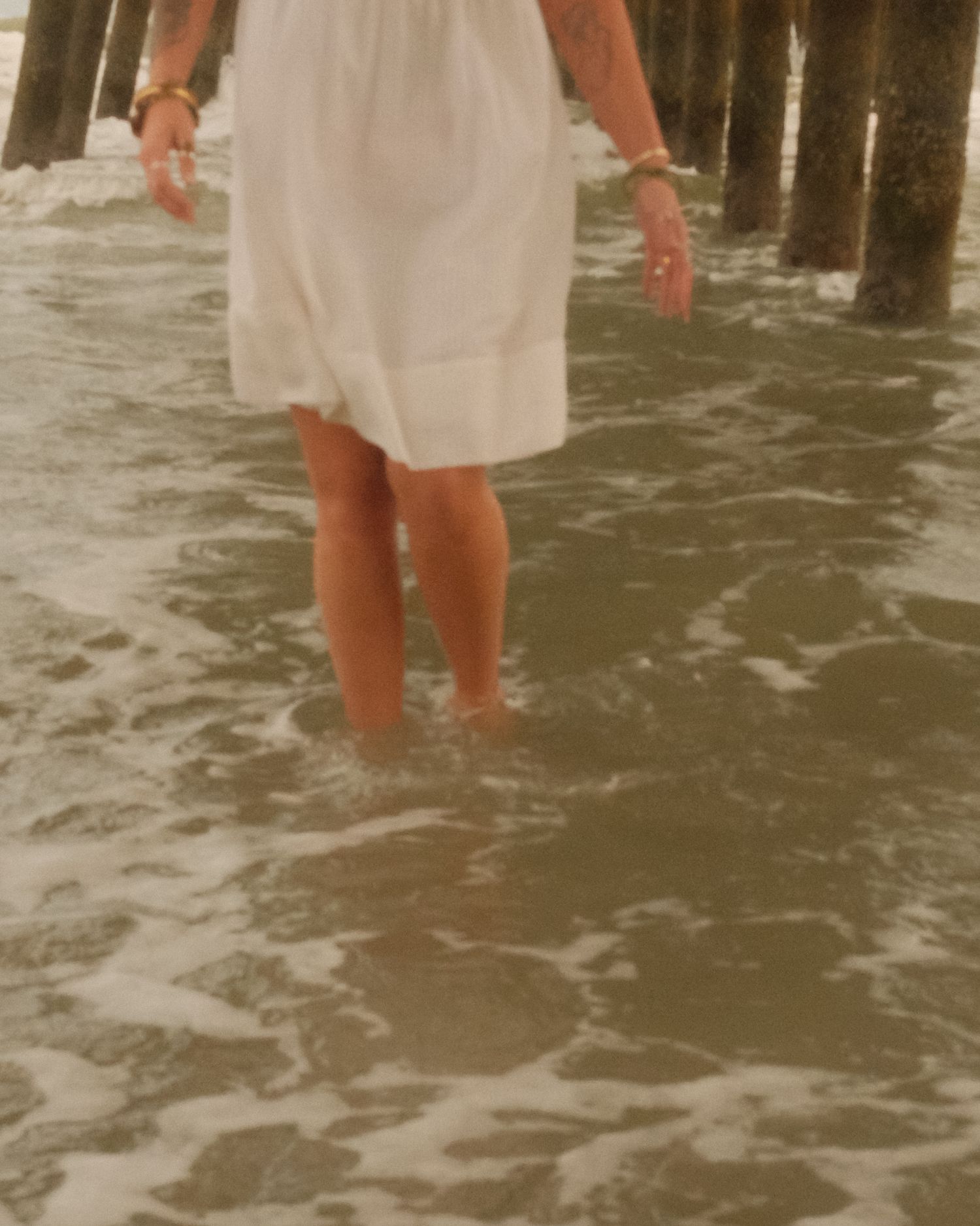 Person in white dress standing ankle-deep in ocean waves near wooden pier posts during sunset.
