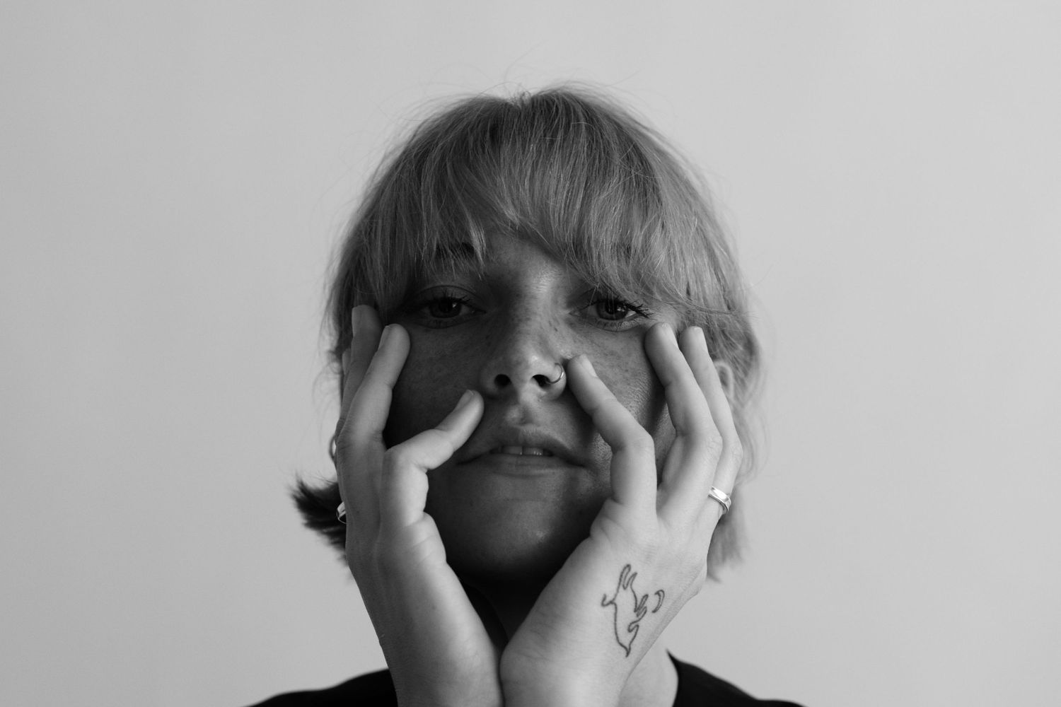 Black and white close-up photograph of someone covering their face with hands while showing a floral tattoo.