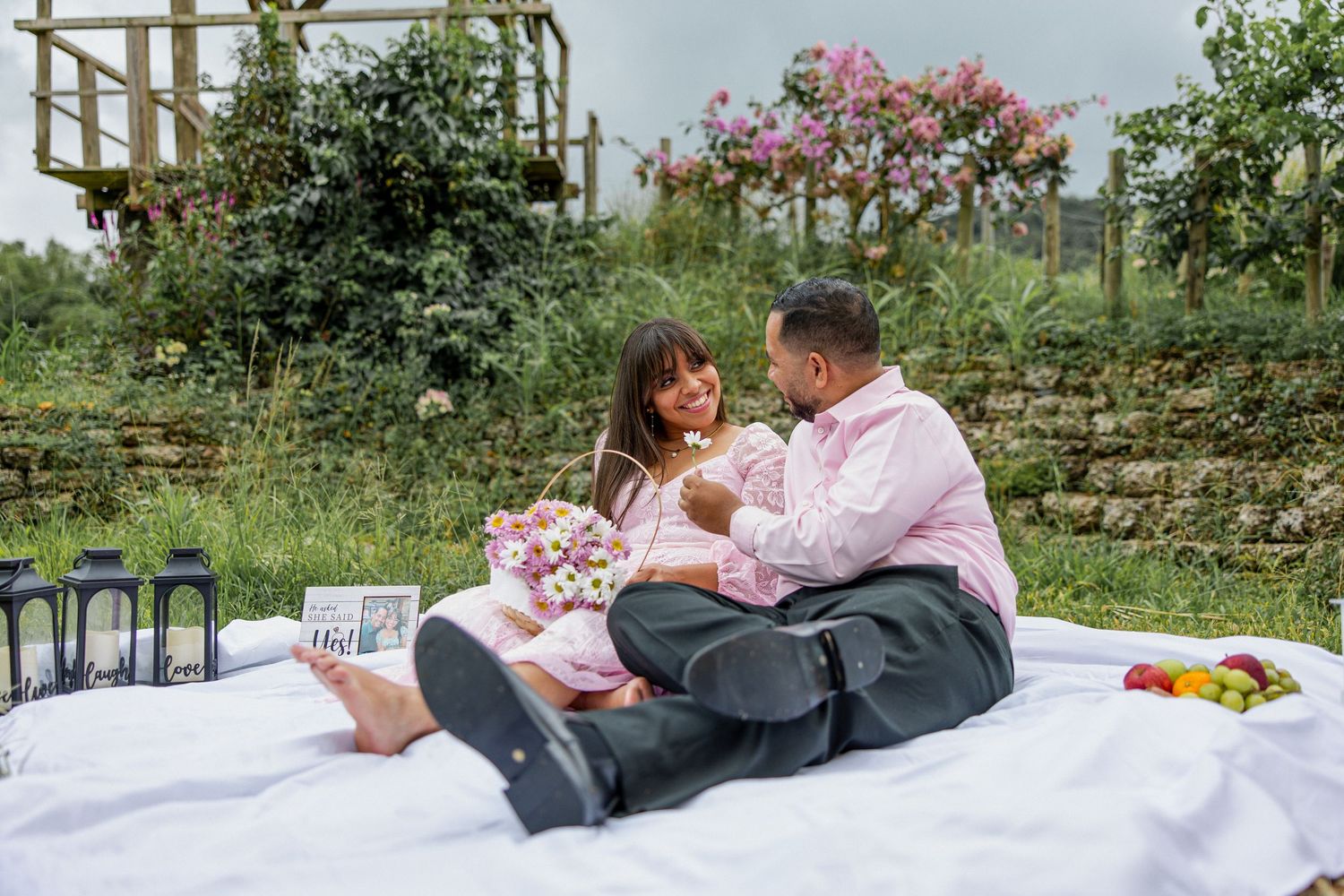 A couple enjoys a romantic picnic with wine and flowers on a white blanket.